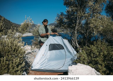 A man sets up a tourist tent among rocks and trees on the seashore. A young tourist guy puts up a small tent on a rocky hiking trail - Powered by Shutterstock