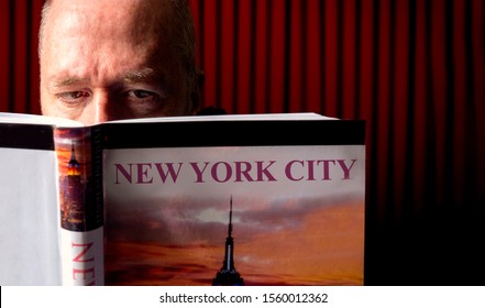 Man In Shadowy Area Of Library Reading Travel Guide Book About New York.