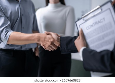 A man shakes hands with another man while holding a contract. A woman is standing behind them. The contract is on a clipboard