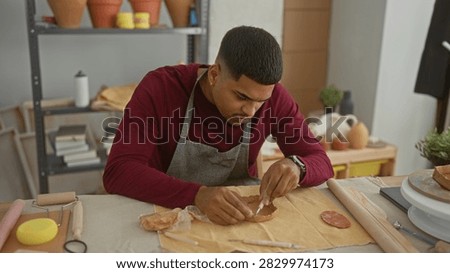 Man shaping clay with hands in studio workshop, wearing apron, leaning over table with sponge brush rollingpin and pottery wheel nearby; quiet concentration.