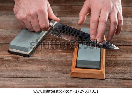 Man sharpening a Japanese knife with a whetstone on a wooden table. Beautiful wavy pattern of Damascus steel blade.