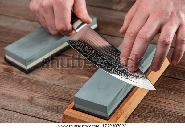 Man sharpens a Gyuto\
knife using a whetstone on a rustic wooden table. Japanese knife\
with Damascus steel.
