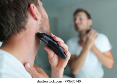 Man shaving beard using electric trimmer shaver. Male beauty grooming concept. Home lifestyle young person looking at bathroom mirror trimming hair on neck.