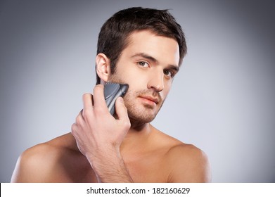 Man shaving. Handsome young man shaving his face with electric shaver and looking at camera while standing isolated on grey background