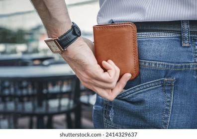 The man in the shirt pulls out a brown leather wallet from the back pocket of his jeans. The street background is out of focus and neutral, focusing on the main object