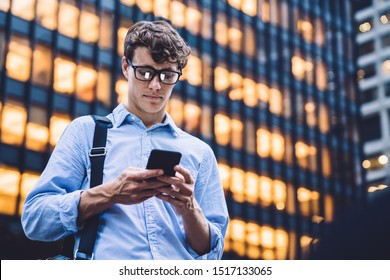 Man in shirt with rolled sleeves and glasses with bag texting on phone standing on background of lighted high building - Powered by Shutterstock