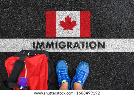 Man in shoes with bag standing next to line with word IMMIGRATION and flag of Canada on asphalt road