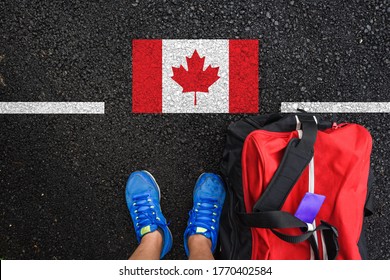 A Man With A Shoes And Travel Bag Is Standing On Asphalt Next To Flag Of Canada And Border