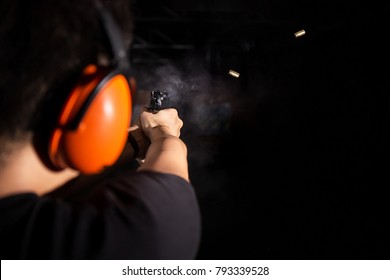 Man Shooting Pistol Gun, Extreme Sport, Fire Bullet, And Wear Orange Ear Cover, Safety Glasses, Equipment In Professional And Safety Shooting Range With Smoke And Bullet Shell On Black Background