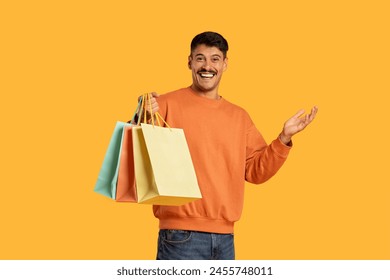 A man with short brown hair and a happy expression is standing outdoors holding multiple shopping bags in his hands. He is wearing a grey t-shirt and jeans, engaging with the camera with a smile. - Powered by Shutterstock