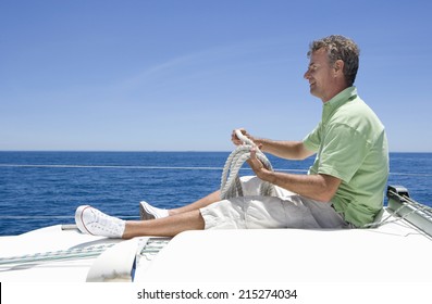 Man In Shorts And Green Polo Shirt Sitting On Deck Of Sailing Boat Out To Sea, Holding Rope, Smiling, Profile