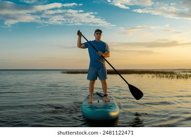 A man in shorts and a T-shirt swims on a lake on a paddle board against the backdrop of a sunset sky with clouds. Horizontal photo. - Powered by Shutterstock