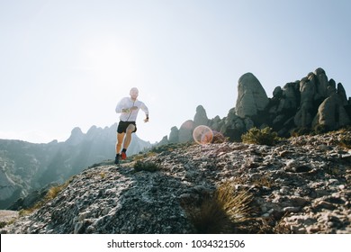Man in shorts and wind jacket runs along rock or cliff on mountain path, during ultra marathon trail race or training on warm sunny day, with beautiful scenery and landscape