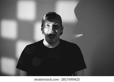 Man with shouting expression looking at camera. Portrait of young caucasian male wearing baseball cap with dramatic shadow lighting. Artistic photography of human emotion and reaction.