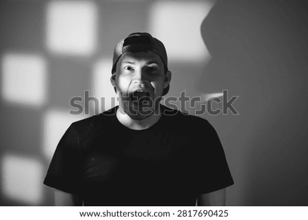 Man with shouting expression looking at camera. Portrait of young caucasian male wearing baseball cap with dramatic shadow lighting. Artistic photography of human emotion and reaction.