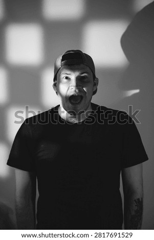 Man with shouting expression looking at camera. Portrait of young caucasian male wearing baseball cap with dramatic shadow lighting. Artistic photography of human emotion and reaction.
