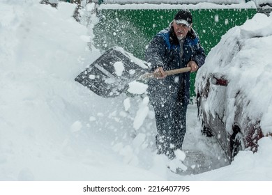 A Man Is Shoveling Snow From The Area Near The House. Nearby Is A Car Covered With Snow. It Is Snowing Outside. A Snowy Winter In Europe. The Global Problems Of Climate Change.