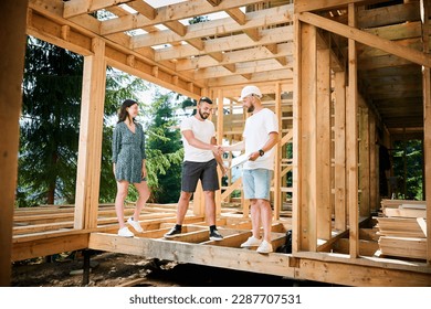 Man showcases to couple the process of constructing wooden framed house near the forest. Happy men firmly grasp each other's hands in a gesture of mutual respect. Concept of reaching mutual agreement.