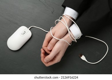 Man showing hands tied with computer mouse cable at black table, top view. Internet addiction