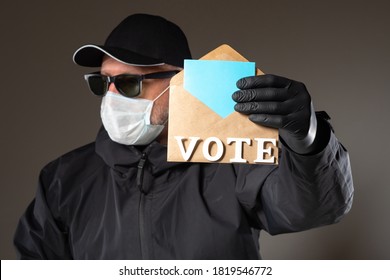 Man Shows An Envelope With Words VOTE. Elections Sign. Voter In Black Jacket And Medical Mask. Concept - Voting During COVID-19. Voting By Mail Over Coronavirus. Conducting Elections Through Postal. 