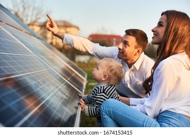 Man shows his family the solar panels on the plot near the house during a warm day. Young woman with a kid and a man in the sun rays look at the solar panels. - Shutterstock ID 1674153415