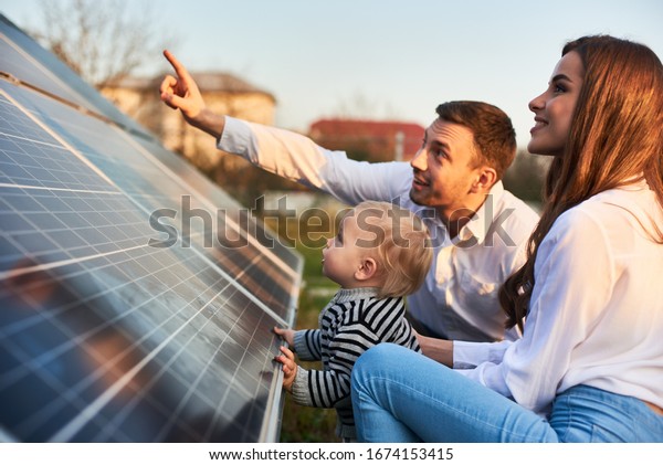 Man shows his family the solar\
panels on the plot near the house during a warm day. Young woman\
with a kid and a man in the sun rays look at the solar\
panels.