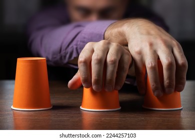 Man shows shell game of thimbles with coin, dark background. Concept deception, sleight hand