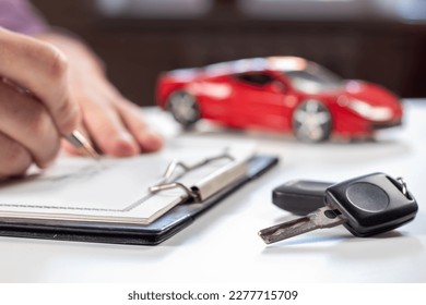 Man signing car insurance document or lease paper. Writing signature on contract or agreement. Buying or selling new or used vehicle. Car keys on table on red car background. Warranty or guarantee.