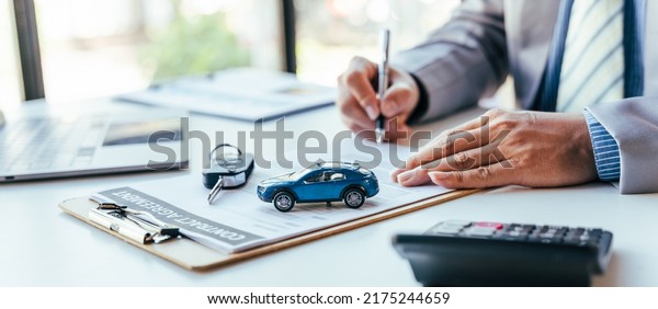 Man signing car insurance document or lease\
paper. Writing signature on contract or agreement. Buying or\
selling new or used vehicle. Car keys on table. Warranty or\
guarantee. Customer or\
salesman.