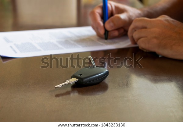 Man signing rental car\
or paper insurance document. Written signature in contract or\
agreement. Buy or sell new or used vehicles. The car keys on the\
table. selective focus