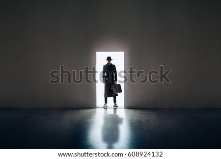 Man silhouette in hat and raincoat standing in the light of opening door in dark room
