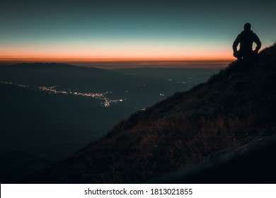 Man Silhouette Standing On The Mountain And Looking Down At The City Lit Up At Night