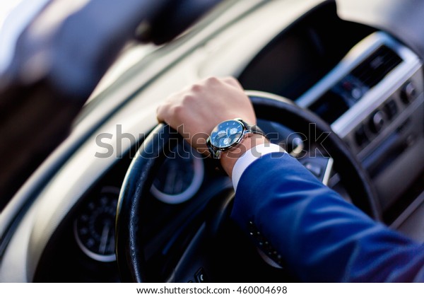 A man in a simple blue\
suit sitting behind the wheel of an expensive car and holds the\
steering wheel