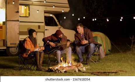 Man singing a song on guitar for his friends around camp fire in a cold night of autumn in the mountains. Retro camper van. Light bulbs. - Powered by Shutterstock