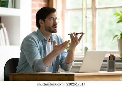 Man Sit At Desk With Laptop Holds In Hand Cell Phone Talks To Client On Speakerphone, Make Formal Conversation Remotely Use Mobile Phone, Send Voice Message. Virtual Assistance, Advanced User Concept