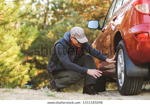 Man sit down near red car checking\
wheels. Elderly man in tourist clothes in\
forest.