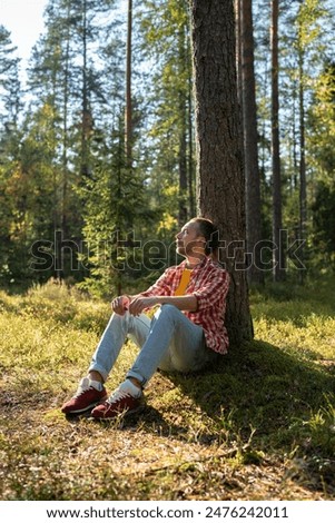 Man sits by tree in forest. Middle aged male resting in woodland, secluded with nature, basking in warm bright daylight. Introverted guy gains strength, restores mental balance. Slow life concept