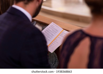 
A Man Sits In The Church And Sings Along To The Church Paper. The Church Sheet, Music Book Is Open. You Can See The Person From Behind. He Has A Beard And A Suit.