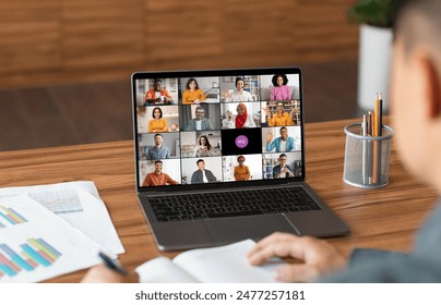 A man sits at a desk in a home office and uses a laptop to participate in a video conference meeting. The laptop screen displays a grid of video windows featuring a diverse group of people - Powered by Shutterstock