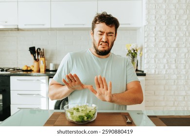 A man sits at a dining table in a stylish kitchen, expressing displeasure while rejecting a bowl of fresh salad in front of him. The atmosphere is bright and modern.