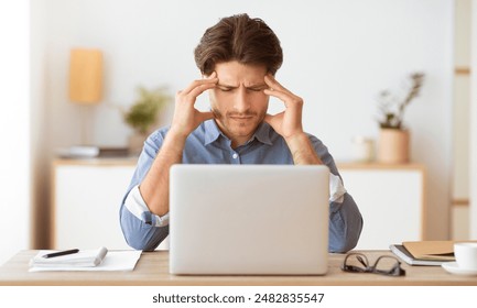 A man sits at his desk with a laptop open, looking stressed and holding his head. - Powered by Shutterstock