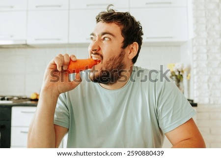 A man sits at a kitchen table, taking a big bite of a crunchy carrot. The bright kitchen is filled with sunlight, and flowers are visible in the background.