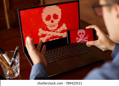 Man Sits Near Laptop With Phone Blocked And Encrypted By Ransomware Spyware Asking For Money. Laptop And Smartphone Infected By Virus. Scary Red Skull Crossbones On Screen. Cyber Security Concept.