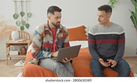 Man sits on bed using laptop while partner holds smartphone beside him in a bedroom with orange blanket and houseplants; contemplation support.