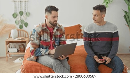 Man sits on bed using laptop while partner holds smartphone beside him in a bedroom with orange blanket and houseplants; contemplation support.