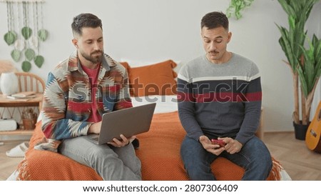 Man sits on bed using laptop while partner holds smartphone beside him in a bedroom with orange blanket and houseplants; contemplation support.