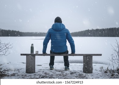 A man sits on a bench on the shore of a frozen lake. Winter. Snow falls. Thermos and a mug are standing nearby. The man is in focus.