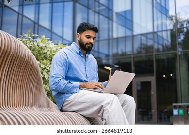 A man sits on a bench, using a laptop outdoors, with a coffee cup in hand, in front of a modern building.