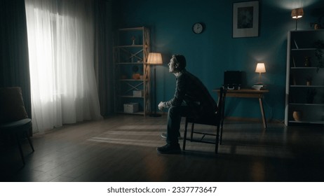 A man sits on a chair in the middle of the room and looks at the light in the window. Side view of a lonely man sitting in a dark room. Hope concept, mental health. - Powered by Shutterstock