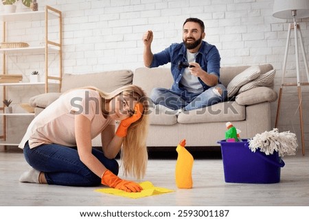 A man sits on the couch, seemingly amused, while a woman kneels on the floor, distressed and cleaning. The atmosphere reflects conflict in their marriage, highlighting domestic abuse.